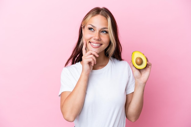 Giovane donna russa che tiene avocado isolato su sfondo rosa pensando a un'idea mentre guarda in alto