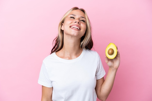 Giovane donna russa che tiene avocado isolato su sfondo rosa ridendo