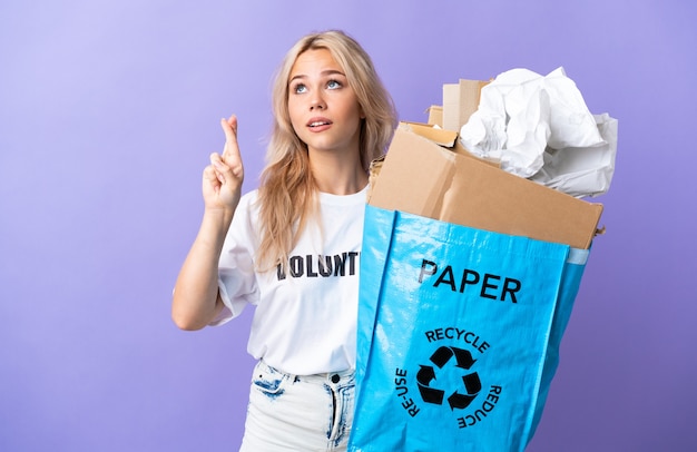 Giovane donna russa che tiene un sacchetto di riciclaggio pieno di carta da riciclare isolato su sfondo viola con le dita che si incrociano e che desiderano il meglio