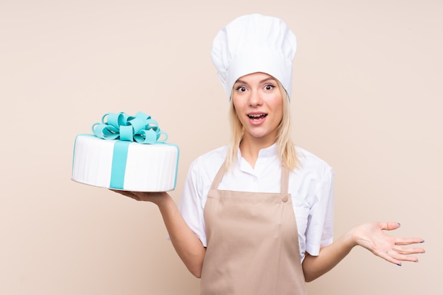 Giovane donna russa con una grande torta sopra la parete isolata con espressione facciale scioccata