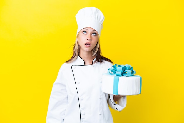 Giovane donna russa del cuoco unico con una grande torta isolata su fondo giallo che osserva in su e con l'espressione sorpresa