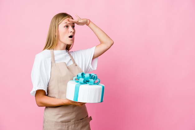 Giovane donna russa del panettiere che tiene una torta deliziosa che guarda lontano mantenendo la mano sulla fronte