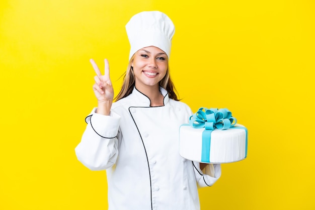 Giovane donna russa del pasticcere con una grande torta isolata su fondo giallo che sorride e che mostra il segno di vittoria