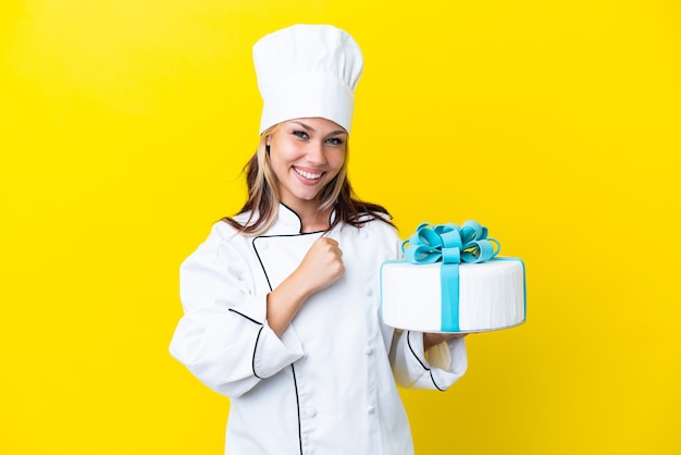 Giovane donna russa del pasticcere con una grande torta isolata su sfondo giallo che celebra una vittoria