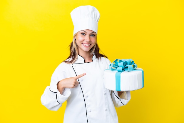 Giovane donna russa del pasticcere con una grande torta isolata su sfondo giallo che indica il lato per presentare un prodotto