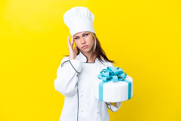 Giovane donna russa del pasticcere con una grande torta isolata su sfondo giallo con mal di testa