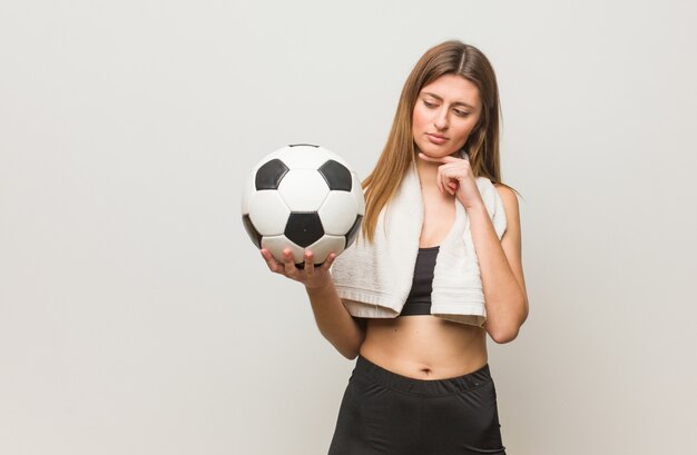 Giovane donna russa di forma fisica che dubita e confusa. Tenendo un pallone da calcio.