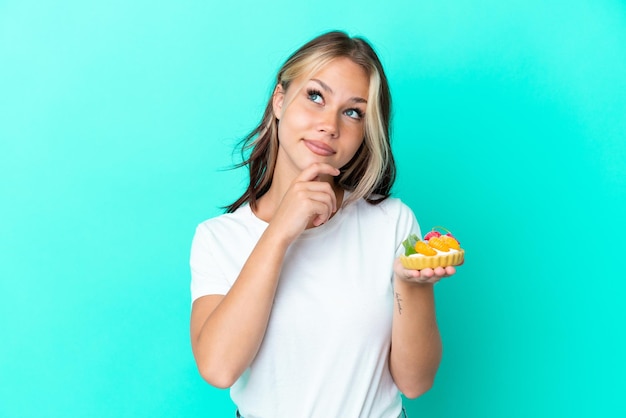 Giovane donna russa in possesso di un dolce di frutta isolato su sfondo blu e alzando lo sguardo