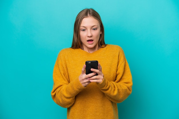 Giovane donna russa isolata su sfondo blu guardando la telecamera mentre si utilizza il cellulare con espressione sorpresa