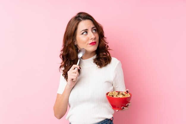 Giovane donna russa sopra fondo rosa isolato che tiene una ciotola di cereali e di pensiero