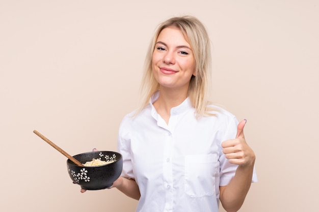 Giovane donna russa sul muro isolato con il pollice in alto perché è successo qualcosa di buono mentre si tiene una scodella di noodles con le bacchette