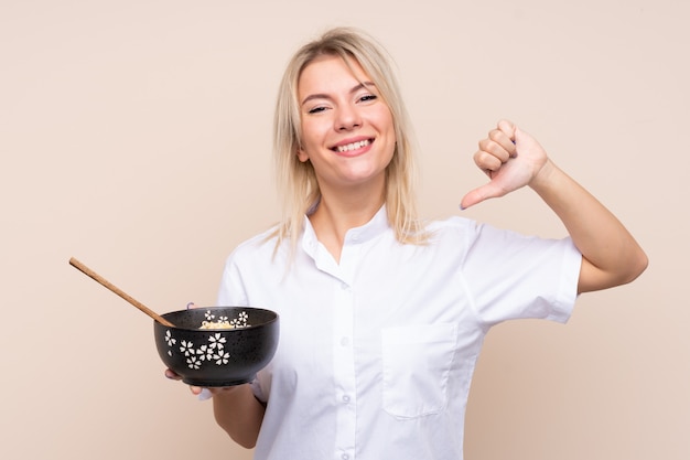 Giovane donna russa sul muro isolato orgoglioso e soddisfatto di sé mentre si tiene una ciotola di noodles con le bacchette