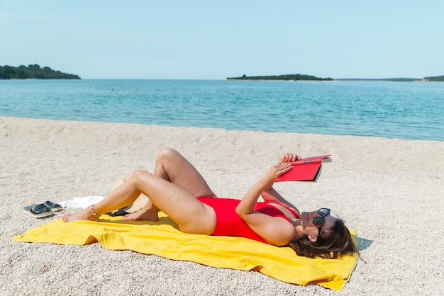 Giovane donna sbalorditiva sexy in costume da bagno rosso che legge il libro sull'ora legale della spiaggia del mare