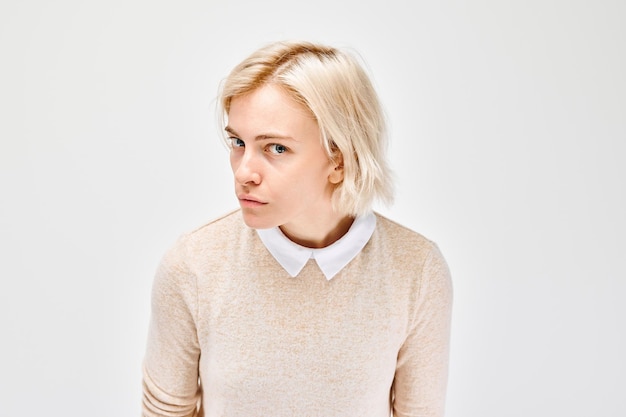 Giovane donna scettica con i capelli biondi corti che indossa un maglione beige su una camicia a collare che guarda di lato contro uno sfondo bianco