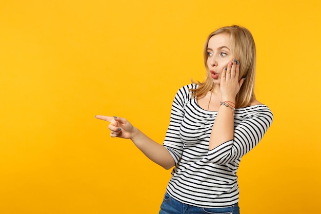 Giovane donna scioccata in abiti a righe che punta il dito indice da parte mettendo la mano sulla guancia isolata su sfondo giallo arancione in studio. Persone sincere emozioni, concetto di stile di vita. Mock up spazio di copia.