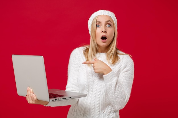 Giovane donna scioccata in cappello maglione bianco isolato su sfondo rosso ritratto in studio. Stile di vita sano della moda, concetto di stagione fredda. Simula lo spazio della copia. Puntare il dito indice sul computer portatile.