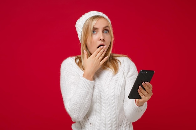 Giovane donna scioccata in maglione bianco, cappello isolato su sfondo rosso. Stile di vita sano della moda, concetto di stagione fredda. Simula lo spazio della copia. Utilizzando il telefono cellulare, digitando un messaggio sms, coprendo la bocca con la mano.