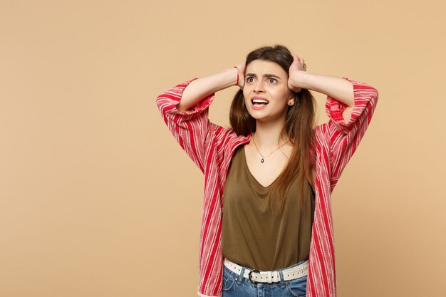 Giovane donna scioccata perplessa in abiti casual che guarda da parte, mettendo le mani sulla testa isolata sul fondo della parete beige pastello in studio. Persone sincere emozioni, concetto di stile di vita. Mock up copia spazio.