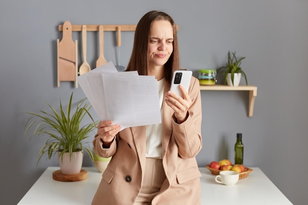 Giovane donna scontenta che risolve le finanze in cucina tenendo un documento cartaceo utilizzando il telefono cellulare con un'espressione perplessa confusa che indossa un abito beige