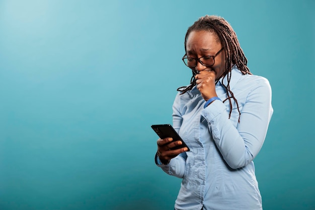 Giovane donna sconvolta che piange dopo aver letto notizie tragiche sul dispositivo smartphone mentre si trova su sfondo blu. Triste persona adulta infelice in lacrime mentre legge un messaggio sfortunato.