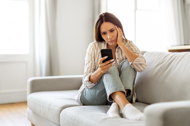 Giovane donna sconvolta seduta sul divano e che guarda il suo smartphone