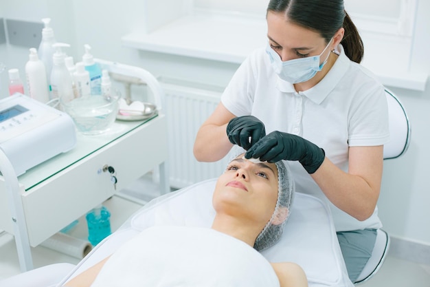 Giovane donna sdraiata su un lettino medico e una signora in maschera medica che si pulisce la fronte con un batuffolo di cotone