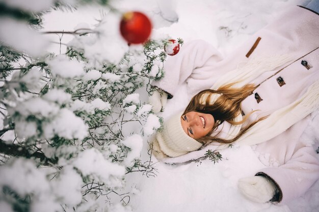 Giovane donna sdraiata sulla neve sotto un abete decorato con palle rosse in una foresta invernale innevata