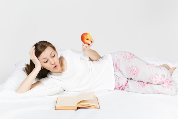 Giovane donna seduta a letto con lenzuolo bianco, cuscino, coperta sul muro bianco