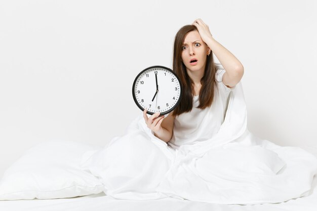 Giovane donna seduta a letto con orologio rotondo, lenzuolo bianco, cuscino, avvolto in una coperta sul muro bianco