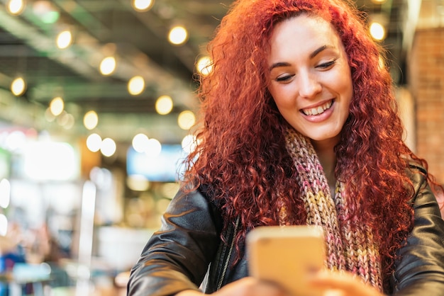 Giovane donna seduta al coperto in un caffè urbano alla moda che scrive con il suo telefono cellulare.