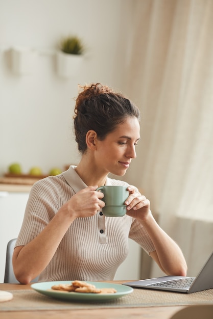 Giovane donna seduta al tavolo e bere il caffè mentre si utilizza il laptop a casa