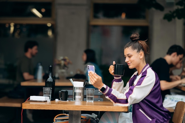 Giovane donna seduta all'aperto caffè e bere caffè tenendo smartphone