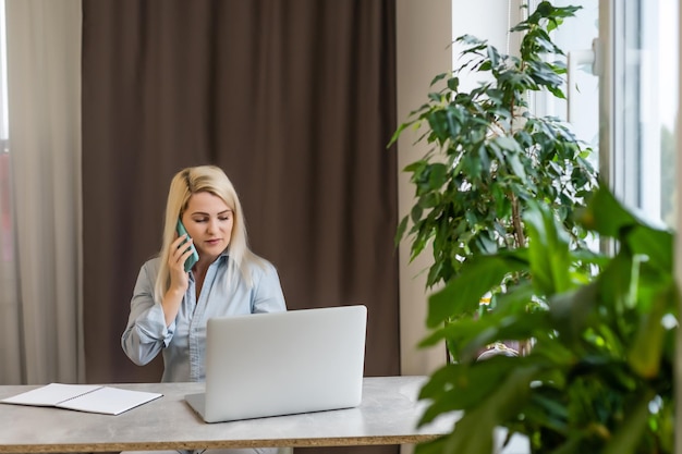 Giovane donna seduta alla sua scrivania davanti a un laptop grigio mentre sogna ad occhi aperti bei ricordi o futuro in un moderno ufficio luminoso con ampia finestra