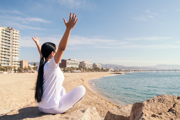 Giovane donna seduta con le braccia alzate che fa yoga sulla spiaggia, concetto di spiritualità e relax