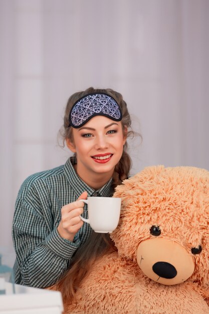 Giovane donna seduta con tazza bianca e orso giocattolo sorridente nella maschera di sonno