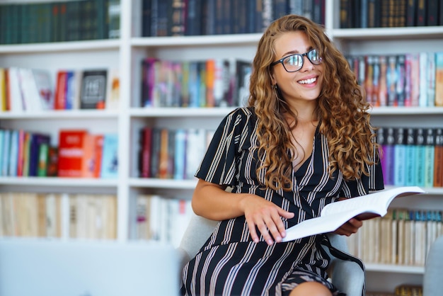 Giovane donna seduta con un libro portatile in biblioteca e prendere appunti di apprendimento Concetto di educazione