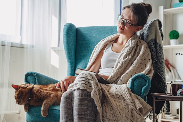 Giovane donna seduta in una poltrona accogliente, con una calda coperta, insieme a un gatto domestico