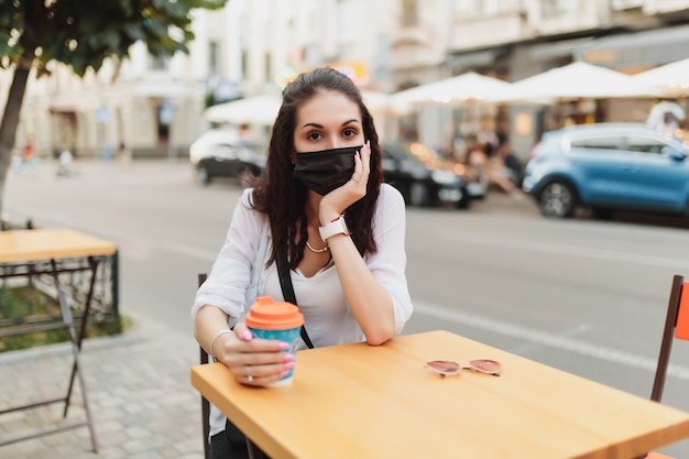 Giovane donna seduta per strada in una maschera con una tazza di caffè. Foto di alta qualità