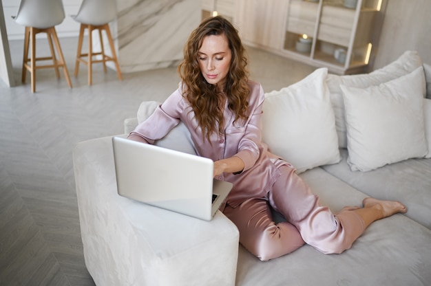 Giovane donna seduta su un divano bianco in stile scandinavo soggiorno cucina interna e lavorando su un computer portatile.