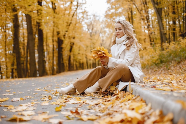 Giovane donna seduta su un marciapiede nella foresta d'autunno. Donna bionda che tiene foglie gialle. Ragazza che indossa cappotto beige e pantaloni marroni.