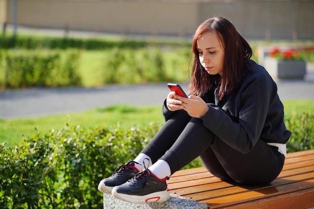 Giovane donna seduta su una panchina del parco guardando il suo cellulare Una donna che utilizza uno smartphone utilizza Internet nel cortile di un edificio residenziale