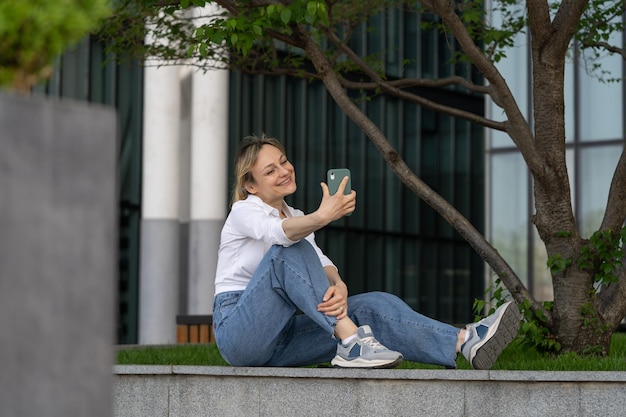 Giovane donna seduta su una panchina sotto l'albero all'aperto che riposa facendo videochiamate online sullo smartphone