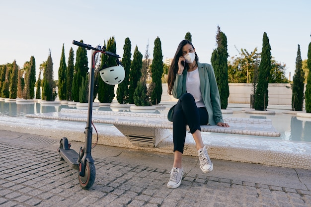 Giovane donna seduta su una panchina utilizzando un telefono cellulare. Donna che indossa la maschera per il viso mentre si utilizza uno scooter elettrico