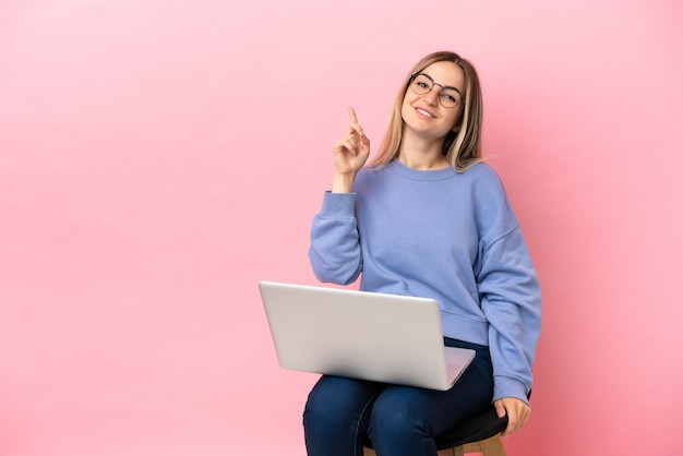 Giovane donna seduta su una sedia con un computer portatile su sfondo rosa isolato che mostra e solleva un dito in segno del meglio