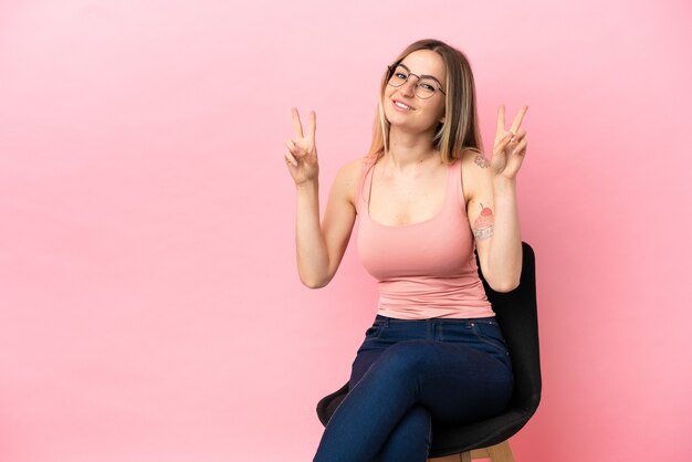 Giovane donna seduta su una sedia su sfondo rosa isolato che mostra il segno della vittoria con entrambe le mani