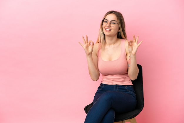 Giovane donna seduta su una sedia su sfondo rosa isolato che mostra un segno ok con le dita