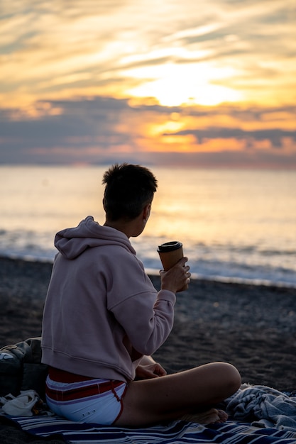 Giovane donna seduta su una spiaggia con una tazza di caffè all'alba o al tramonto