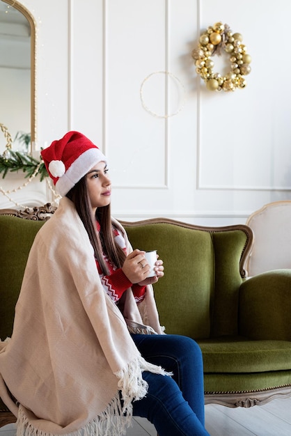 Giovane donna seduta sul divano da sola in un soggiorno decorato per il Natale con una tazza di bevanda calda