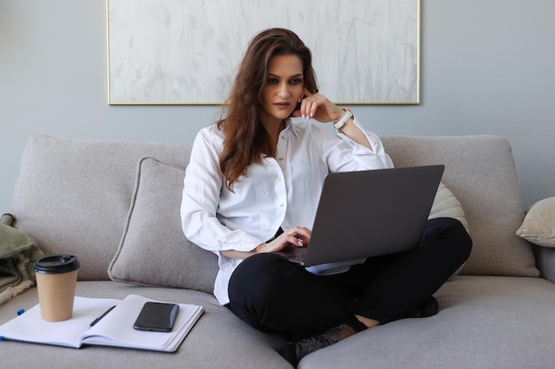 Giovane donna seduta sul divano e lavorando su un laptop a casa