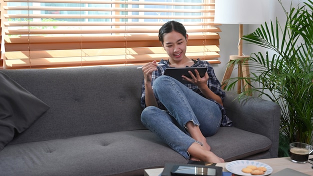 Giovane donna seduta sul divano in soggiorno e utilizzando tablet.